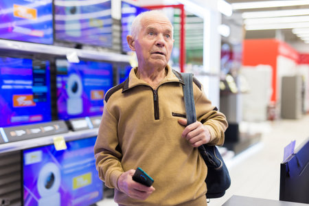 Elderly grayhaired man pensioner looking counter with modern digital televisors in showroom of digital goods storeの写真素材