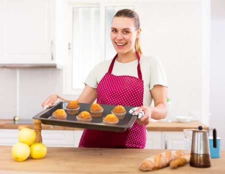 Smiling woman in apron standing in home kitchen with tray of cupcakesの写真素材