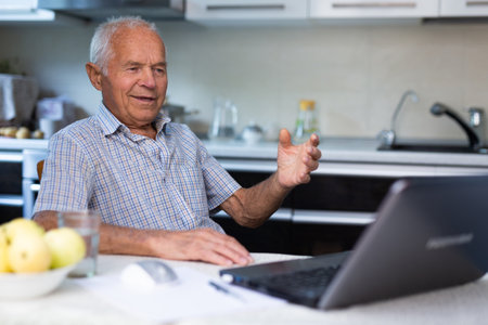 Retiree doing online shopping using laptop while sitting in the kitchen of homeの写真素材