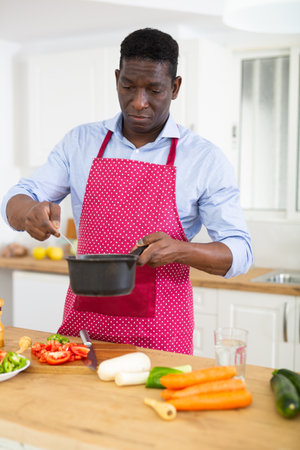 Smiling adult man in red apron preparing vegetable dish in home kitchenの写真素材