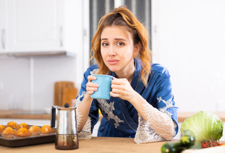 Upset sleepy woman in blue dressing gown drinking coffee in kitchenの写真素材