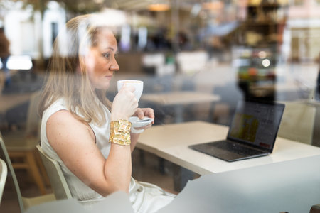 Business lunch - businesswoman sits at table in cafe by window with a laptop and drinks coffee. View through windowの写真素材