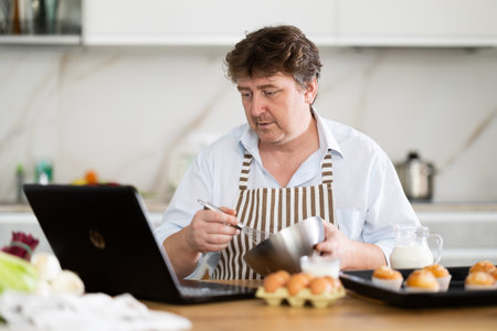 Man in chef hat and apron prepares cupcakes in kitchen according to recipe from the Internet -...