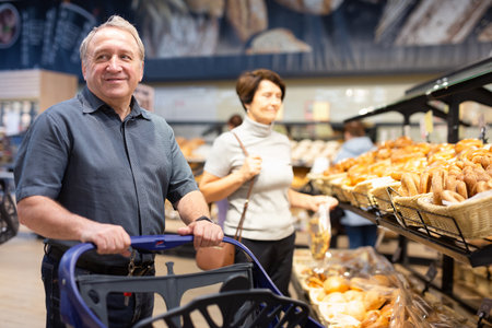 Elderly male shopper with a shopping cart chooses groceries in supermarketの写真素材