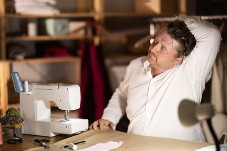 Middle-aged male tailor stretching while sitting next to sewing machine at night