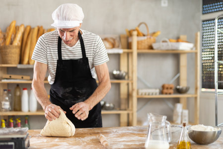 Elderly male baker preparing raw yeast dough on kitchen table in bakery