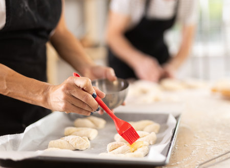 Female hands brushing uncooked dough pieces on baking sheet with oil before baking in oven closeup