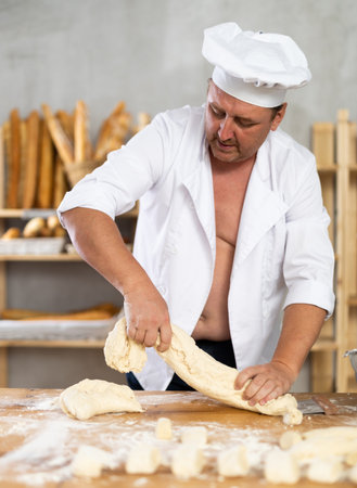 Male baker in unbuttoned uniform prepares dough, kneads it with his handsの写真素材