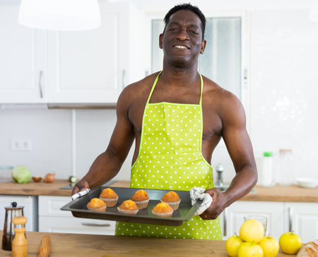 Smiling man wearing yellow apron holding baking tray with muffins in kitchenの写真素材