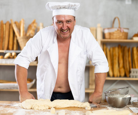 Male baker in unbuttoned uniform prepares dough, kneads it with his hands