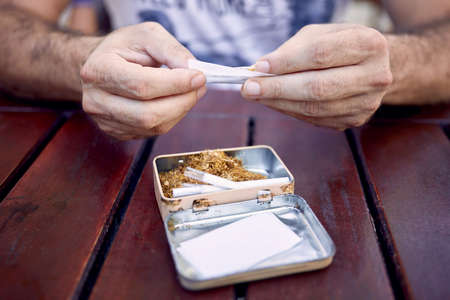 The hands of a caucasian male rolling a cigarette on a wooden table. Cigarette paper, filters, and a tin box full of a tobacco is on the table.の写真素材