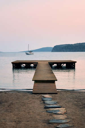A peaceful sunrise scene with pastel colors including sand, stones, jetty, boat, sky, and mountains...