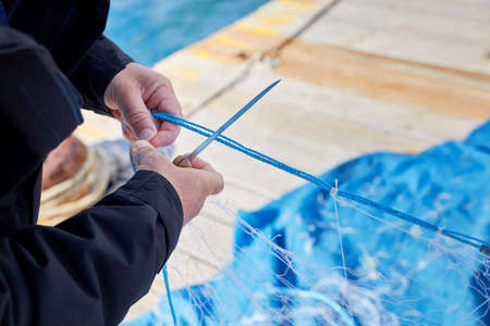 Hands of a fisherman repairing a fishing net with a knife on the harborの写真素材