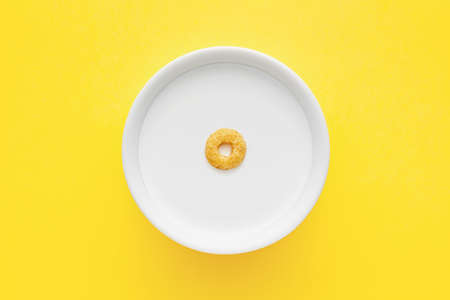 Minimalist view of a single cereal ring floating on a white bowl full of milk on yellow background.の写真素材