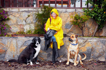 Smiling Caucasian female at her 30s wearing a yellow raincoat and sitting on a stone wall in between...