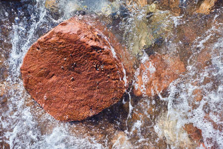 Big red rock in the sea surrounded with colorful rocks and water.