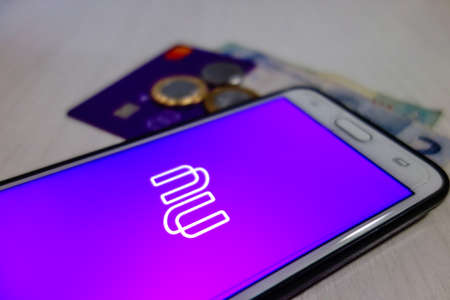 Nubank brazilian fintech logo visible on smartphone screen, with credit card and money on backgroundのeditorial素材