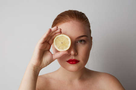 Close-up portrait of pretty young woman with red lips with closed eyes and holding a fresh lemon and posing on the gray wall. Isolated.の写真素材