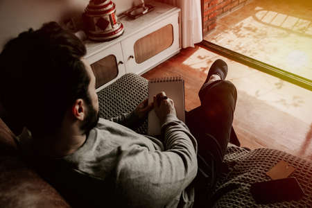Back side picture of young man writing a text in the note book, sitting on the chair at home.