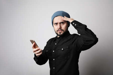 Handsome and bearded hipster wearing stylish hat on head using his smartphone with serious face.