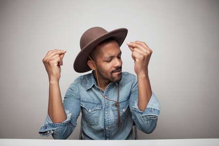 Close-up portrait of young black man wearing denim shirt putting hands up posing against grey wall...