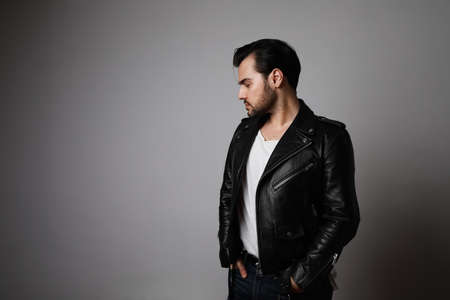 Side shot of brutal man with dark beard, looks aside, wears biker jacket, over blank studio background with free space.の写真素材