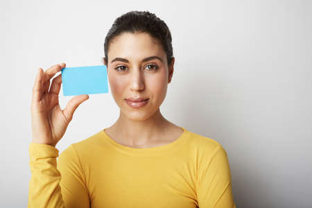 Portrait of a pretty young woman dressed in sweater holding credit card at her face isolated over white background.の写真素材