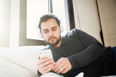 Attractive smart young man sitting on a sofa in the living room and using his smartphone. Relaxing time.の写真素材