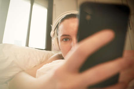 Headshot of young cheerful woman taking a self-portrait and laying at the bed. Space for text.