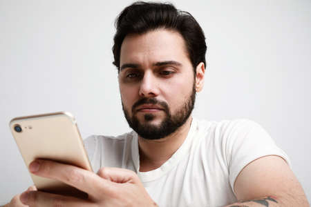 Headshot of attractive young man with a beard using a smartphone and texting while posing over white background.の写真素材