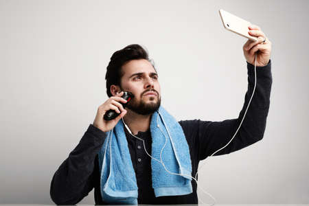 Handsome bearded man trimming his beard with a trimmer and using his smartphone watching video tutorials.の写真素材