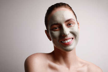 Close-up portrait of happy woman with clay facial mask with cute smile isolated on grey wall. Beauty and skin care.の写真素材