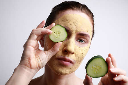Close-up of young woman with a face mask on holding a cucumber slices in front of one eye. Beauty and skincare concept.の写真素材