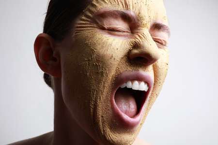 Close-up portrait of young woman with clay mask on her face against light background, space for...