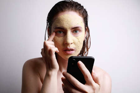 Portrait of young caucasian woman with facial clay mask using her smartphone watching videos. Space...