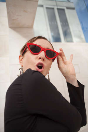 Vertical portrait of happy and cheerful mature woman outdoor, wearing retro red sunglasses with exclamation grimace.の写真素材