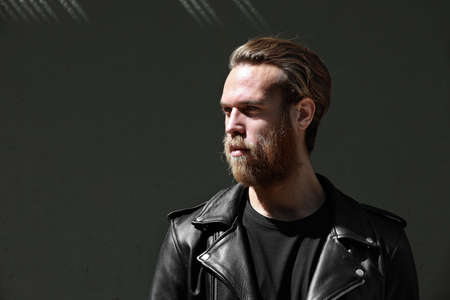 Brutal young man with beard, looks thoughtfully aside, wears biker jacket posing over black background with free space.の写真素材
