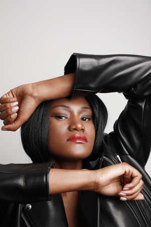 Vertical close-up portrait of young pretty black woman wearing a leather jacket against white wall.