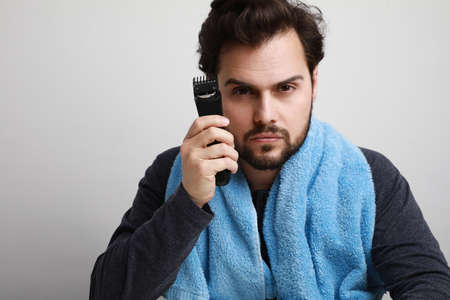 Headshot portrait of handsome young man shaving his beard in bathroom, holding shaver machine. Space for text.の写真素材