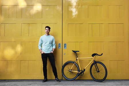 A young stylish hipster caucasian man posing next to his bicycle on the yellow background.