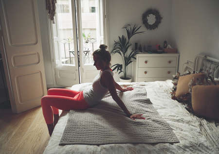 Blonde woman is sitting on the bed. Girl is doing yoga at home. Space for text.の写真素材