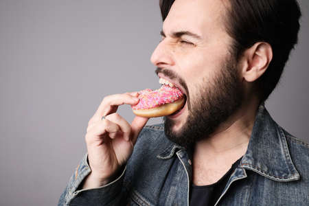 Cropped side portrait of young man biting pink donut piece with pleasure.の写真素材