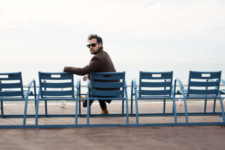Bearded man in coat sitting on the bench against the sea coast.