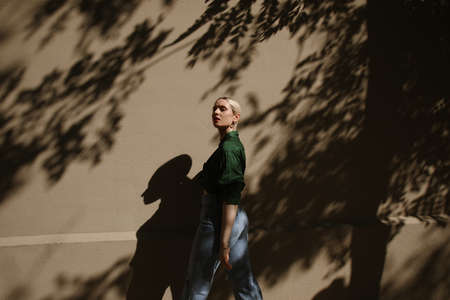 Young blonde woman posing on beige wall with dark shadows looks at the camera.の写真素材