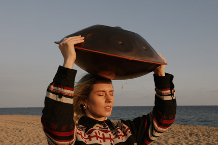 Caucasian woman holds hang drum on head while sitting on the beach.の写真素材