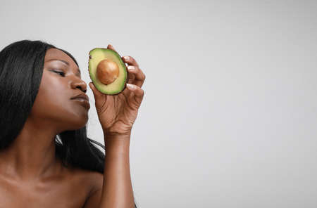 Gorgeous black young woman holds half-part of avocado posing on white wall.の写真素材