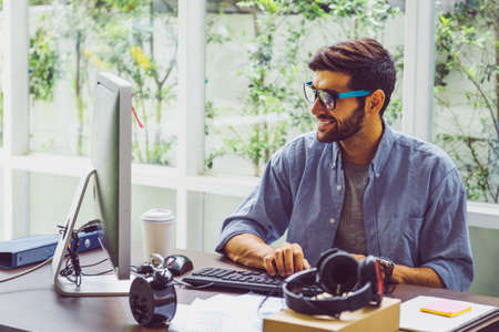 Happy young man, wearing glasses and smiling, as he works on his laptop to get all his business done early in the morningの写真素材