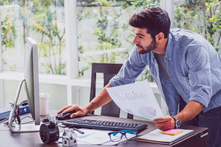 Young bearded businessman stands in the office near table and uses computerの写真素材