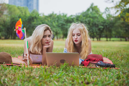 Two young women friends watching social media videos blogs while laying outdoors on green meadow