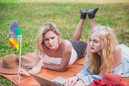 Two young women friends watching social media videos blogs while laying outdoors on green meadowの写真素材